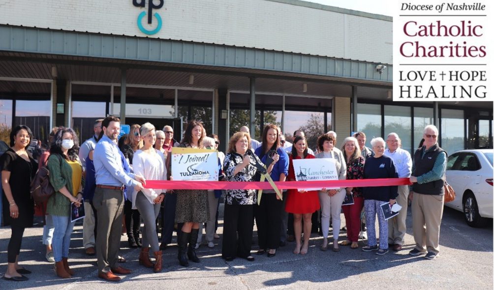Image of group of people standing in front of a gray building for a ribbon cutting. Dioces of Nashville Catholic Charities branded graphic at top right.
