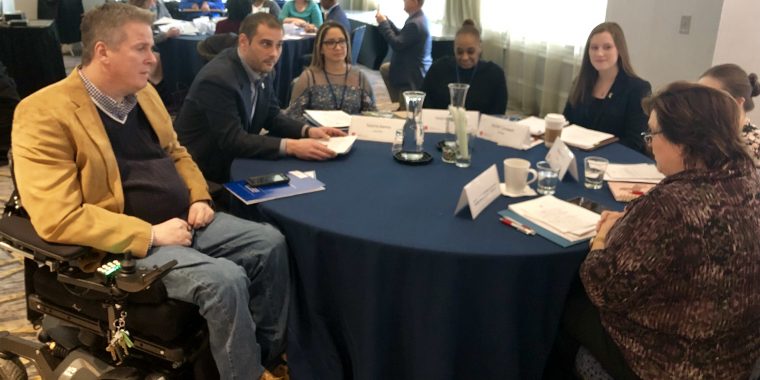 A group of 7 diverse people sitting around a round shaped table with a blue cloth. The man on the left is in a wheelchair.