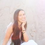 Image of a young adult female wearing a white sundress and sitting in front of a textured wall.