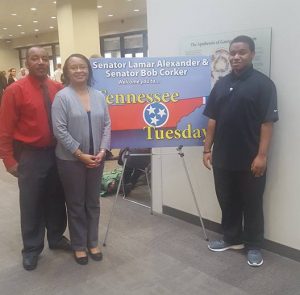 Image of Linda Brown's family (Husband, wife, & son) standing in front of a sign with an image of the state of Tennessee in red with the Tennessee flag. 