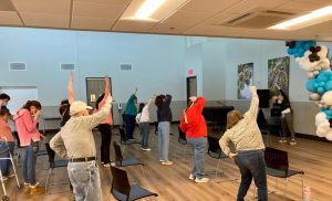Photograph of a group of people standing up and moving their right arm in the air, mirroring the movements of one person standing in front of them. They are inside a room with wooden floors, black chairs, and white, blue, and gray balloons.