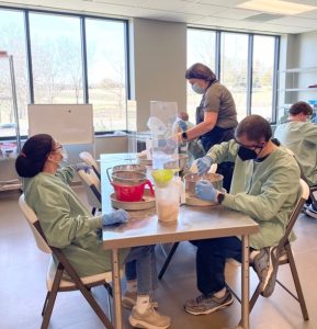 Photograph of people wearing green scrubs, black hair nets, black and blue face masks, and blue medical gloves. Three people are sitting down, and one is standing up, all working with a silver kitchen bowl.