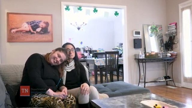 Image of two females sitting on a grey couch in a living room setting. The female on the left is wearing a black top with cheetah print leggings and is lying her head on the right shoulder shoulder of the female on the right who is wearing a black shirt, tan pants, a white scarf, and glasses.