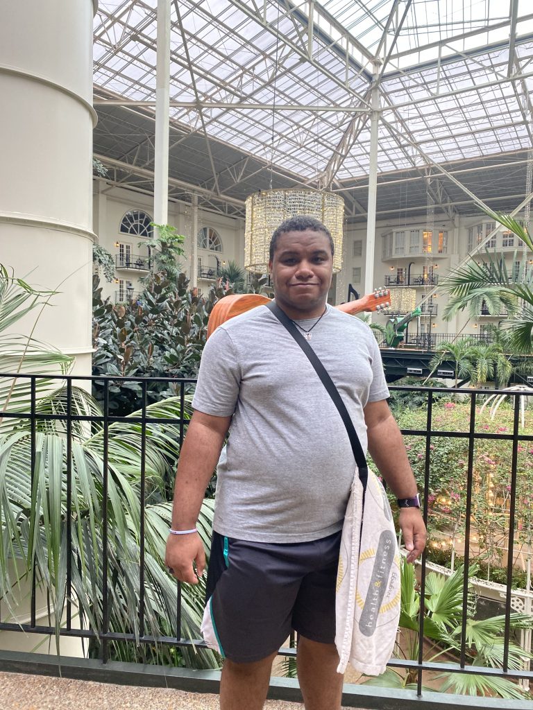Image of a male wearing a gray t-shirt and dark color shorts with a bag strapped across his body. He is standing in front of indoor greenery.