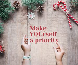 Woodgrain background image with pine cones, snowflakes and peppermints hanging from garland on the top of the image. At the bottom of the image are two hand holding text "Make YOUrself a ppriority."