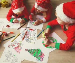 Image of three children wearing red white and green long sleeve tops with red and white Santa hats lying on the floor coloring sheets of paper with markers and colored pencils.