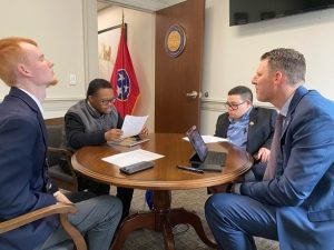 Image of four men in blue suits sitting around a round table with papers on top in an office with a Tennessee flag. 