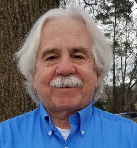 Image of a man with platinum gray medium length hair and a mustache wearing a blue button down shirt with a white t-shirt peeking out at the top. The man is in a wooded area. Image is of Mark G. Friedman, Ph.D.