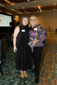 Image of two women standing side by side wearing nametags. The female on the left has dark blonde hair and is wearing a black maxi dress with brown shoes. The female on the left is wearing a colorful top with black pants and black boots. She is holding a book in her left hand. They both are in front of a large projection screen in a large room.