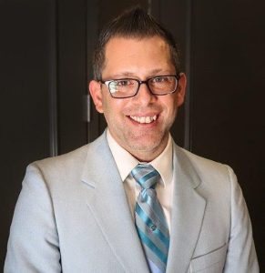 image of Milton wearing a light gray suit jacket, a white shirt and light blue tie with dark rimmed glasses. He has a big smile on his face. 
