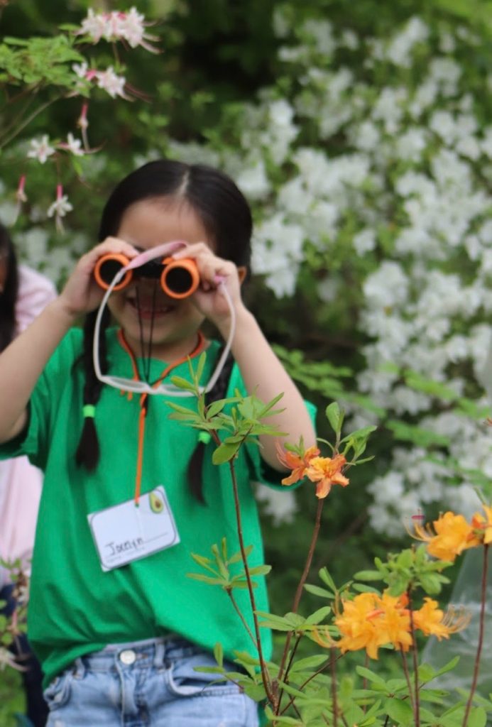 A child wearing a green shirt and blue jeans stands outdoors among blooming flowers and greenery, holding orange binoculars up to their eyes. The child has two braided pigtails with green hair ties and wears a name tag labeled "Jasmine" with an avocado illustration. In the foreground, there are vibrant yellow-orange flowers, while the background features white blossoms and lush foliage.