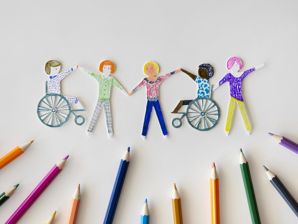 Paper cutouts of children, including two using wheelchairs, holding hands in a row above a semicircle of colorful pencils on a white background, symbolizing inclusion and creativity.