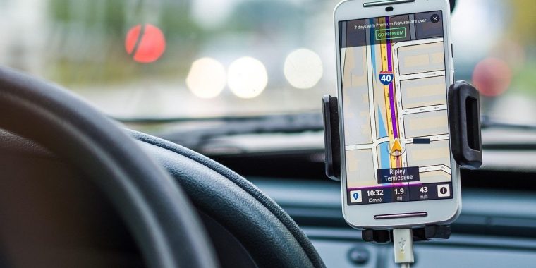 A close-up view from the driver's seat inside a car, showing a smartphone mounted on the dashboard. The phone displays a GPS navigation app with a map and route highlighted in purple and yellow, directing the driver along Interstate 40 as shown by a red, white and blue highway symbol to "Ripley Tennessee," that is shown at the bottom of the phone screen in a dark rectuangular box. The screen shows directions, distance remaining (1.9 mi), estimated time (3 minutes), and current speed (43 m/h). The steering wheel is partially visible on the left, and blurred traffic lights and cars can be seen through the windshield in the background.