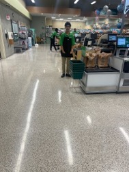 A Youth wearing a green polo shirt, khaki pants a black apron and black shoes working at a grocery store, standing beside a register. The face of the youth has been blurred to maintain privacy. 