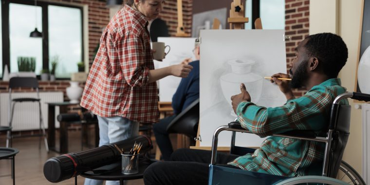 Teacher explaining illustration process to paralyzed student with disability working at painting project during artistic lesson in creativity studio. Diverse team sketching vase using graphic pencil.