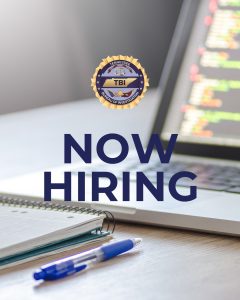 A Tennessee Bureau of Investigation (TBI) badge displayed above bold text that reads “NOW HIRING.” The background shows a laptop with colorful code on the screen, an open notebook, and a blue pen on a desk, suggesting a professional work setting.