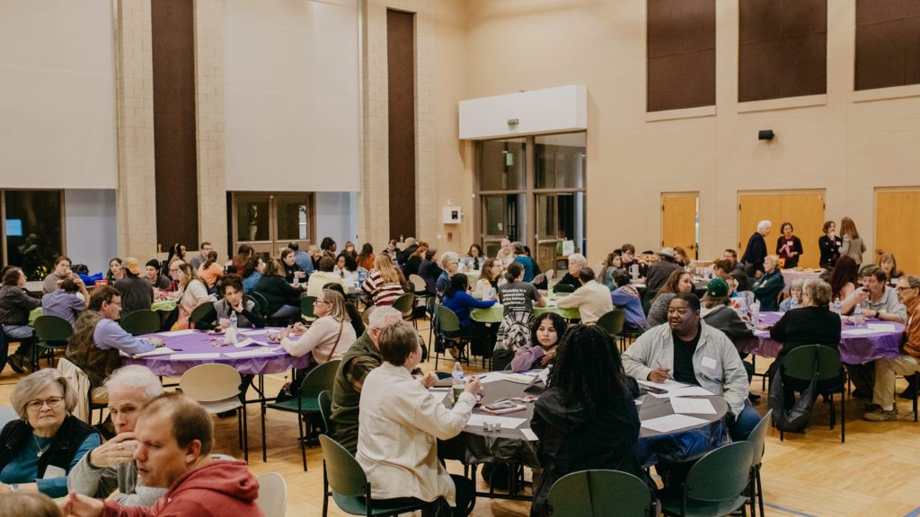 A large community event held in a spacious hall with high ceilings and tall windows. Multiple round tables are arranged across the room, each covered with purple or gray tablecloths and surrounded by chairs. Groups of people are seated at the tables, engaged in conversation and writing on paper. The background shows beige walls, wooden doors, and a bright, well-lit environment with overhead lights.