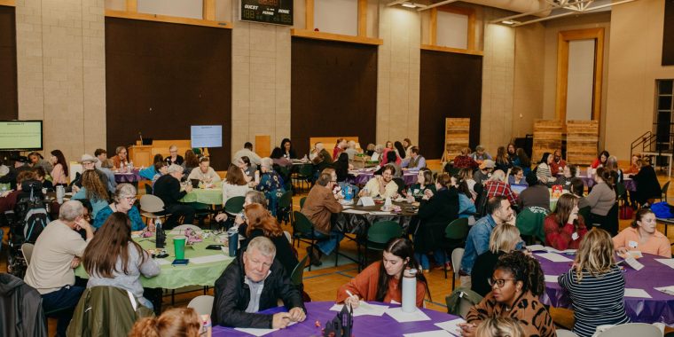 A wide view of a community gathering in a gymnasium-style hall with high ceilings and exposed beams. Numerous round tables covered with purple and green tablecloths fill the space, with attendees seated and participating in discussions. Papers, pens, and water bottles are visible on the tables. Large dark panels line the walls, and two screens are positioned at the back, displaying content for the event.