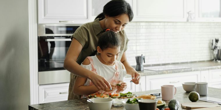 A modern kitchen with white cabinets and a marble countertop. Two people are standing at the counter preparing food together. The counter is filled with fresh ingredients including sliced vegetables, avocados, and bowls of various items. A pink mug and a cutting board are also visible on the counter. Stainless steel appliances are in the background.