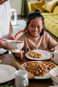 A dining table set for breakfast with white plates, mugs, and a wooden spoon. On the table are several golden-brown waffles arranged on a plate. A person is serving strawberries from a white bowl to another person seated at the table. In the background, there is a green sofa with yellow cushions and a bright, cozy atmosphere.