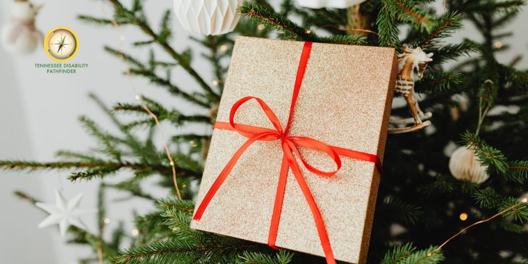A glittery gold gift box tied with a bright red ribbon hangs on a decorated Christmas tree. The tree is adorned with white paper ornaments and warm string lights. The Tennessee Disability Pathfinder logo appears in the upper left corner.