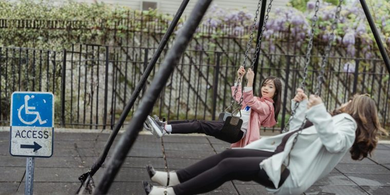 Two people on playground swings are moving forward at the same time. The swings are suspended from a metal frame over a rubber safety surface. A blue sign with a wheelchair symbol and the word “Access” and an arrow pointing right is posted near the swing area. Behind the swings is a black metal fence, greenery, and purple flowering vines.