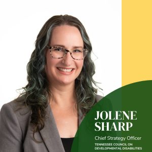 A professional portrait-style image showing an individual from the chest up against a plain white background. The person has long, wavy hair that extends past the shoulders with darker tones and lighter highlights. They are wearing a gray blazer over a dark top. On the right side of the image, a large graphic element features a curved dark-green shape with white text that reads “Jolene Sharp, Chief Strategy Officer, Tennessee Council on Developmental Disabilities.” A vertical yellow bar runs along the upper right edge, adding contrast to the design. The overall layout combines a clean headshot with branded graphic elements suitable for organizational or professional use.