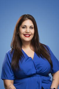 A professional headshot of a woman with long dark brown hair wearing a blue blouse. She is smiling and facing the camera against a soft blue gradient background. She is wearing small hoop earrings and red lipstick, with her hands resting at her sides.