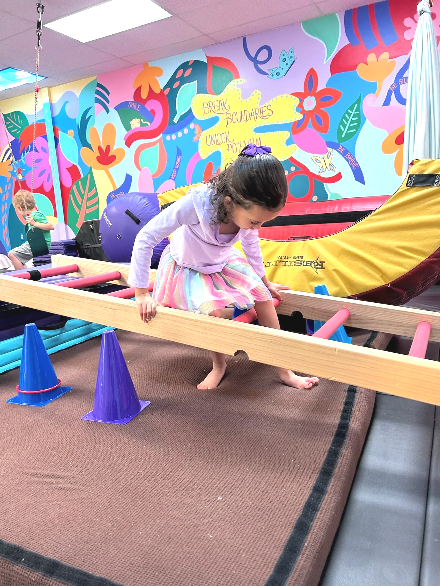 A young child wearing a purple top and a colorful rainbow skirt carefully steps over low wooden balance beams set on cones inside a sensory gym. The child is barefoot and focused while navigating the obstacle. The room features soft mats, foam equipment, and a bright mural on the wall with flowers, leaves, and encouraging words including “Break Boundaries” and “Unlock Potential.” Another child is visible in the background using hanging sensory equipment.