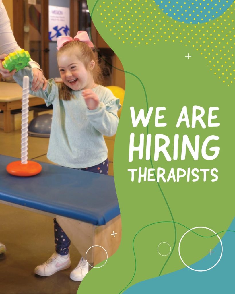 A young child with Down syndrome smiles while participating in a therapy activity at a clinic. The child wears a light blue long-sleeve shirt, patterned leggings, white sneakers, and a large pink bow. She holds a green toy while stacking or interacting with a vertical peg activity on a therapy table. A therapist’s hand is partially visible assisting. Therapy equipment and a treatment room are visible in the background. On the right side of the image, green graphic text reads “We Are Hiring Therapists.”