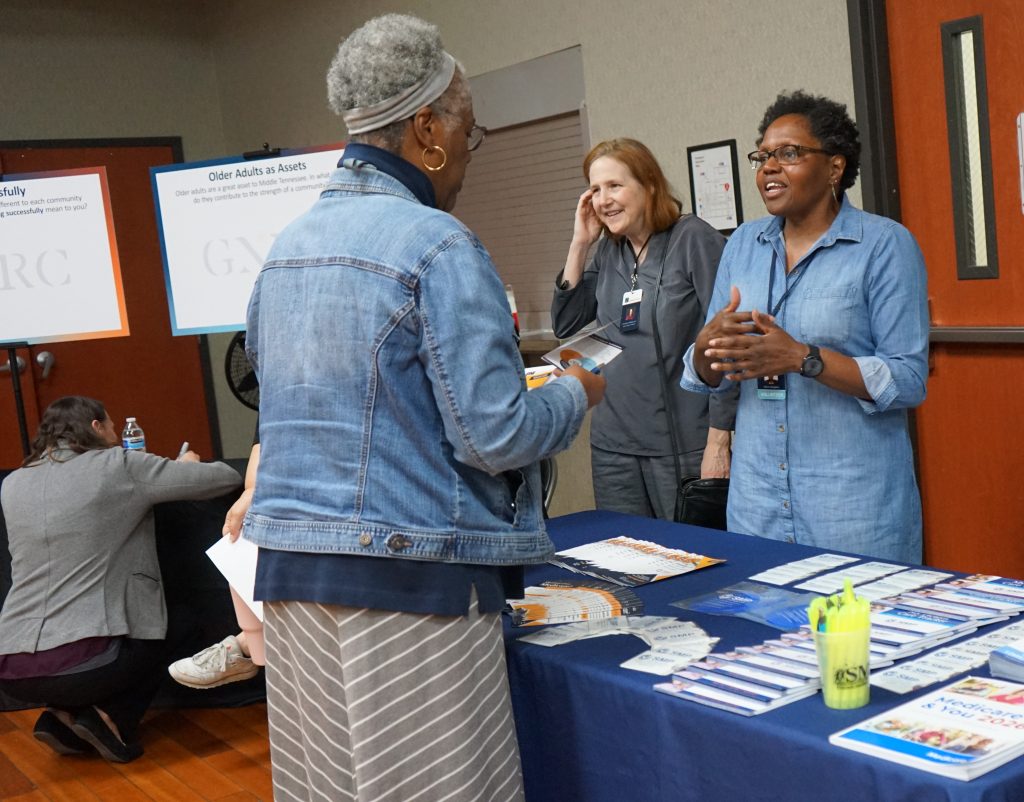 A community outreach and information-sharing event taking place indoors at a resource table. In the foreground, an older Black woman with short gray hair stands facing two women behind a table covered in informational brochures, flyers, pens, and name tags. She wears a light denim jacket over a navy shirt and a gray-and-white striped skirt while holding papers in her hands.One of the women behind the table is a Black woman with short natural hair and glasses, wearing a light denim button-up shirt and a volunteer badge. She gestures with her hands while speaking warmly and attentively to the attendee. Beside her stands a white woman with shoulder-length red hair wearing a gray outfit and name badge, smiling as she listens to the conversation. The table is neatly arranged with stacks of resource materials, including healthcare and community information pamphlets, along with bright yellow highlighters in a clear container. In the background, posters mounted on easels display community engagement topics, including one titled “Older Adults as Assets.” Another attendee is seated near the wall writing or filling out paperwork. The room has wood flooring, beige walls, and a welcoming, collaborative atmosphere focused on community resources, aging, and support services.