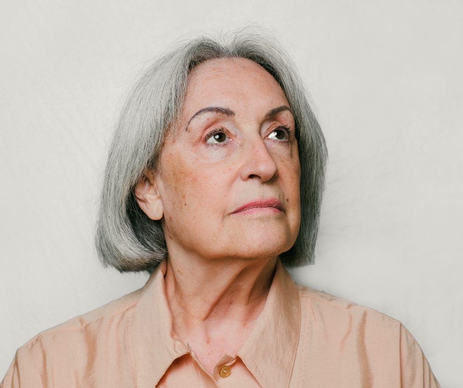 Portrait of an older woman with shoulder-length gray hair styled in a smooth bob, standing against a soft light gray background. She is wearing a light beige button-up blouse with a collar. Her expression is calm, reflective, and slightly serious as she gazes upward and off to the side rather than directly at the camera. Fine details in her face, including soft wrinkles and natural skin texture, are visible, adding realism and warmth to the image. The lighting is soft and even, creating a gentle and thoughtful mood. The composition centers on her face and upper shoulders, giving the portrait an intimate and contemplative feel often associated with themes of aging, memory, or reflection.