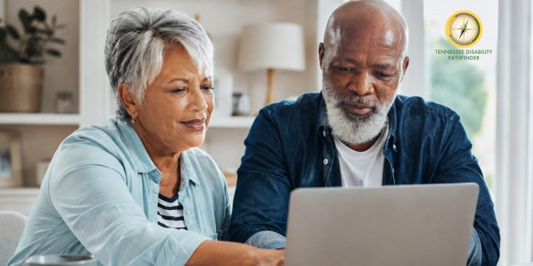 n older Black couple sits side by side at a wooden table in a bright, warmly lit home. The woman, wearing a light blue shirt over a striped top, points toward a laptop screen while the man, wearing a dark blue button-up shirt, leans in and looks closely at the screen. Papers, a notebook, and a coffee mug are on the table, suggesting they are reviewing information together. The image conveys aging adults navigating resources, making plans, and seeking support in a calm, collaborative setting.