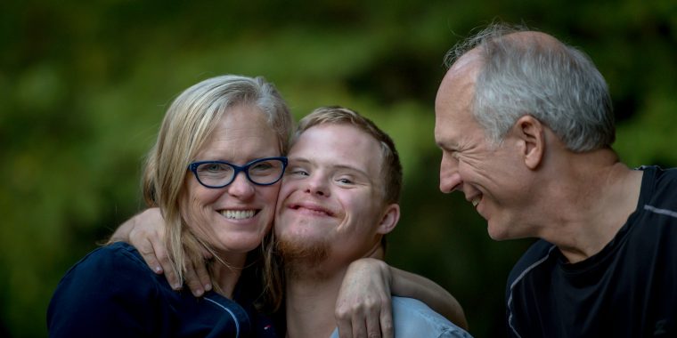 Outdoor portrait of a smiling family sharing an affectionate moment in front of a softly blurred green background filled with trees and foliage. In the center is a young white man with Down syndrome, short light brown hair, and a small goatee. He is smiling warmly while wrapping his arms around the shoulders of the two adults beside him. On the left is a white woman with shoulder-length blonde hair wearing bright blue glasses and a navy-blue top. She smiles directly at the camera while leaning closely into the young man. On the right is an older white man with short gray hair wearing a black athletic shirt. He is turned toward the young man with a wide smile, appearing joyful and engaged in the moment. The trio’s close physical connection and relaxed expressions create a warm feeling of love, family support, and togetherness.