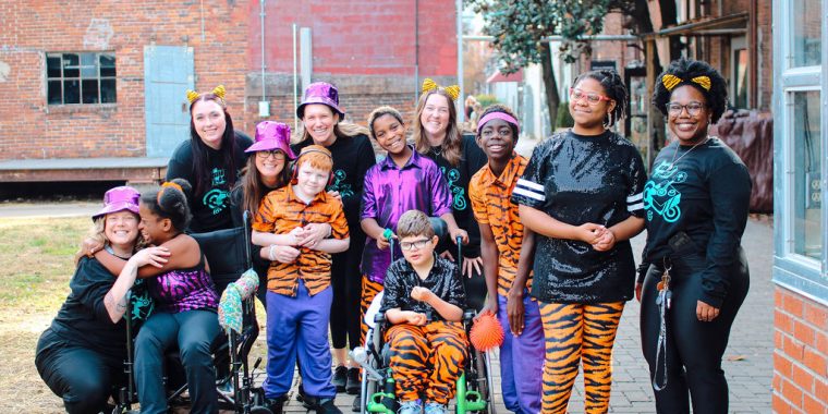 A group of dancers and volunteers pose together before taking the stage for a MOVE Inclusive Dance recital. They are wearing a mixture of orange tiger print and metallic purple costumes as they smile for the camera.