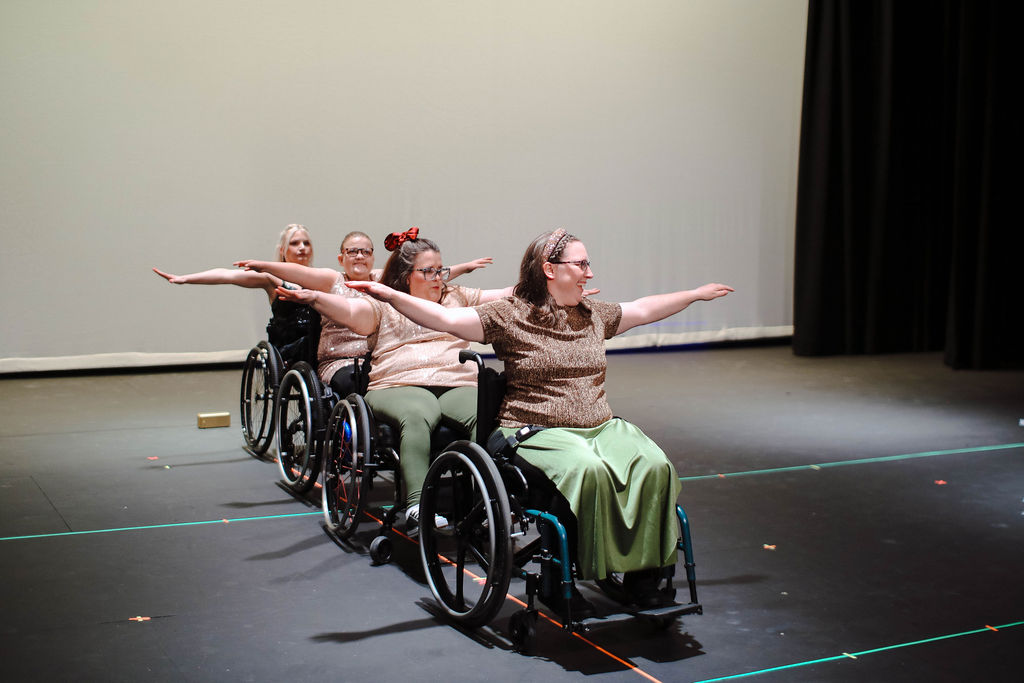 A group of dancers in wheelchairs perform together on stage during a MOVE Inclusive Dance recital. They are wearing a mixture of black, gold, and olive green costumes and are posed in a straight line with their arms extended out.