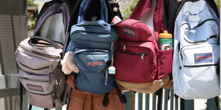 Close-up photograph of four JanSport backpacks hanging side by side outdoors in a school-like setting. The backpacks vary in color, including light lavender, muted blue, deep red with a tan bottom, and light blue, with additional darker backpacks partially visible behind them. Several bags feature the recognizable JanSport logo on their front pockets. The blue backpack has decorative keychains attached, including a fluffy pom-pom and small charm. A green, orange, and white sports drink bottle sits in the side pocket of the red backpack. The image evokes back-to-school preparation, school supplies, and students getting ready for a new academic year.