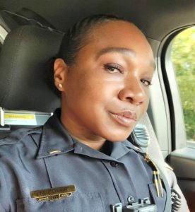 Close-up selfie of a Black woman wearing a dark navy law-enforcement uniform while seated inside a vehicle. Her dark hair is pulled neatly back, and she wears small stud earrings. She looks toward the camera with a calm, composed expression. Her uniform includes a gold nameplate, collar insignia, pens secured in a chest pocket, a body-worn camera partially visible near the bottom of the image, and a shoulder-mounted radio microphone. The vehicle’s dark headrest and interior frame the left side of the photograph, while a window on the right shows softly blurred green trees and daylight outside.