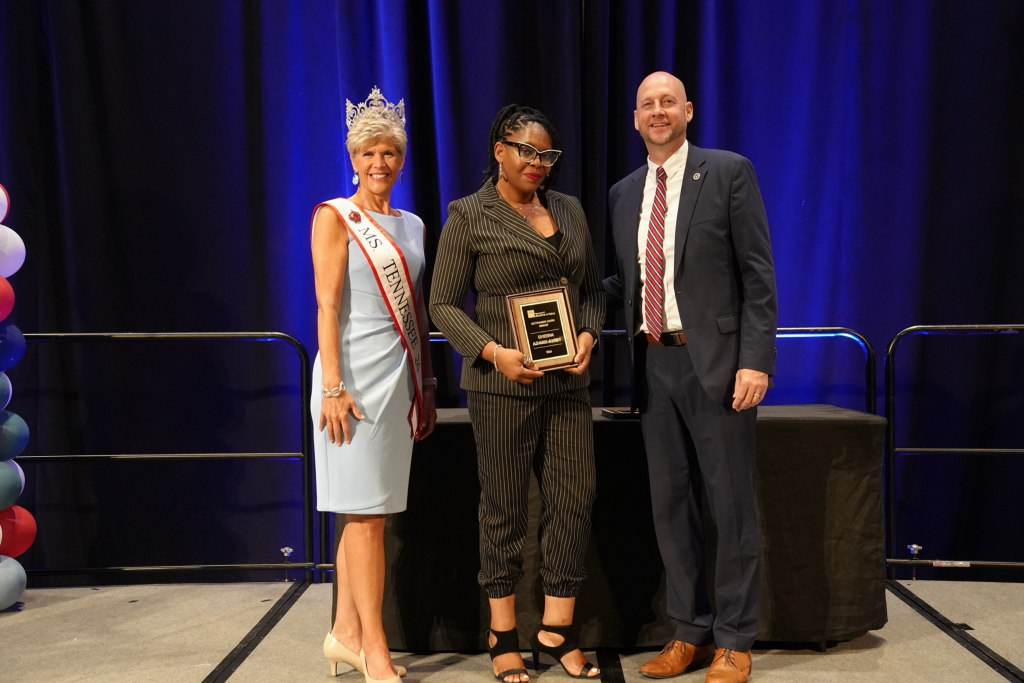 Sheena Adams-Avery stands center stage holding her Reframing Aging Award at the inaugural Tennessee State Conference on Aging awards ceremony. She wears a black pinstripe suit and glasses. Standing to her left is Sandra Elliott, Ms. Tennessee Senior America 2026, wearing a crown and sash. Commissioner Brad Turner stands to Sheena’s right in a navy suit and striped tie. Blue stage lighting and dark curtains provide the backdrop.