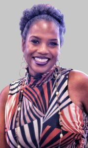 Cristalynne Dupree, Media & Communications Specialist at Tennessee School for the Blind, smiles warmly at the camera. She has natural hair styled in a high puff and wears large hoop earrings and a sleeveless, high-neck top featuring a bold pattern in black, cream, and rust tones. She is photographed against a plain gray background.