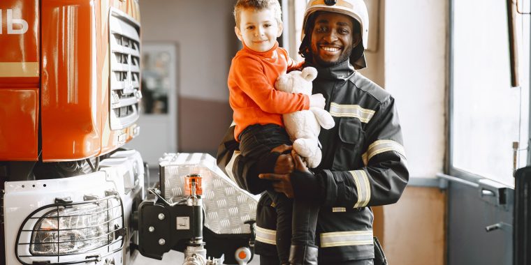 Firefighter and Child: A smiling Black firefighter wearing a white helmet and dark protective uniform holds a young boy dressed in an orange sweater and holding a stuffed animal. They stand beside a large orange fire truck inside a fire station.